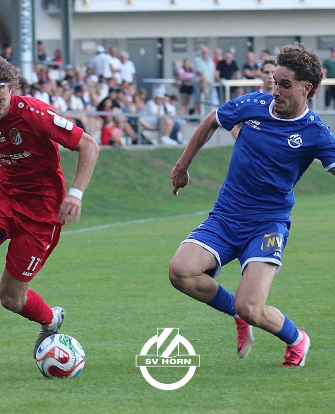 Two soccer players in action on a field, one in red and one in blue, with a crowd watching from the stands.