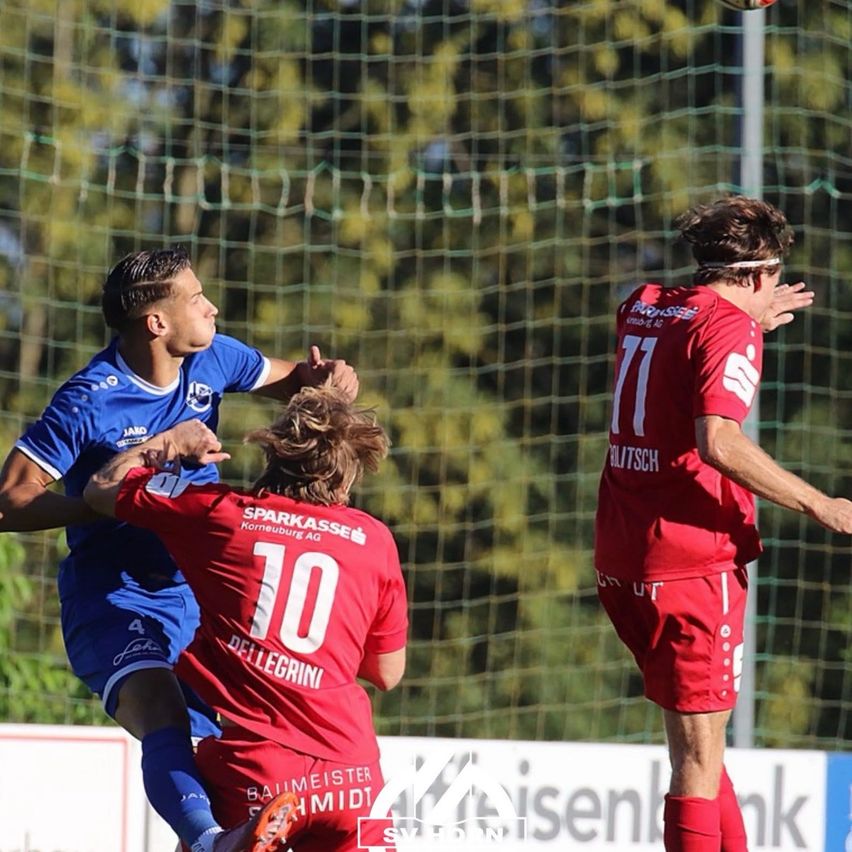 Three soccer players in action, one in blue and two in red, with one player attempting to block a shot.