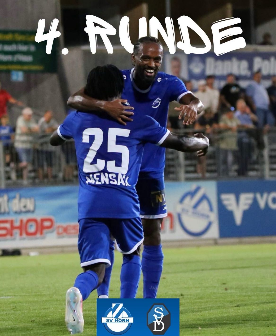 Two soccer players in blue uniforms celebrate on the field with spectators in the background.
