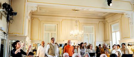 Eine Gruppe Erwachsener in einem reich verzierten Raum mit goldenen Akzenten, einer Laterne und Fenstern. Einige stehen, andere sitzen und einige spielen Instrumente.