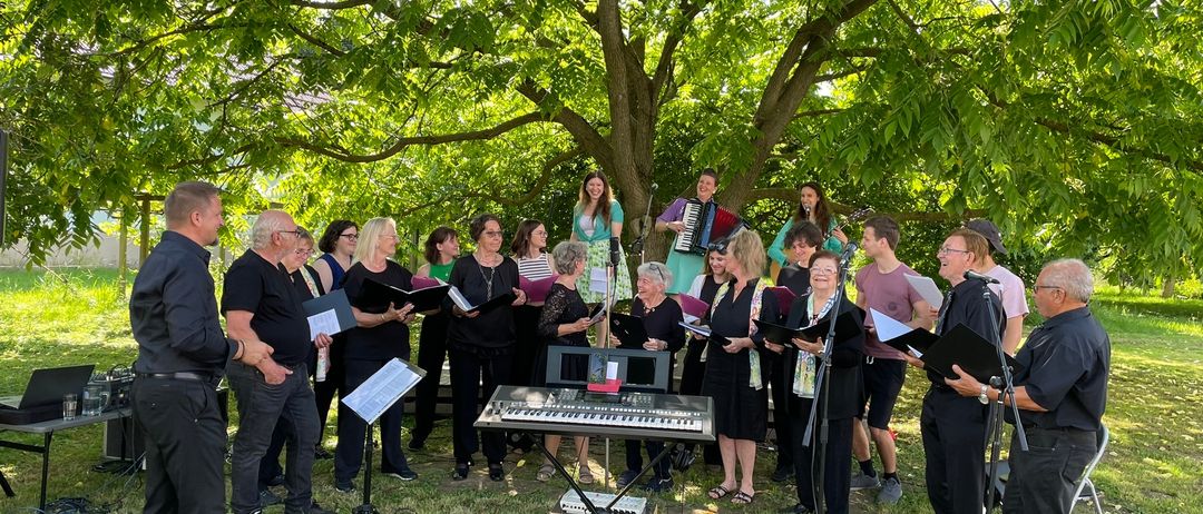 Ein Chor erwachsener Menschen in Schwarz versammelt sich unter einem Baum und singt, während sie Musikblätter halten. Ein Klavier und Mikrofone sind für ihre Aufführung aufgebaut.
