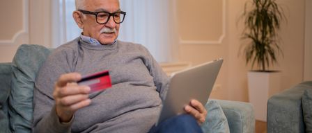Ein älterer Mann mit Brille sitzt auf einem Sofa, hält eine Kreditkarte in der Hand und benutzt einen Laptop.