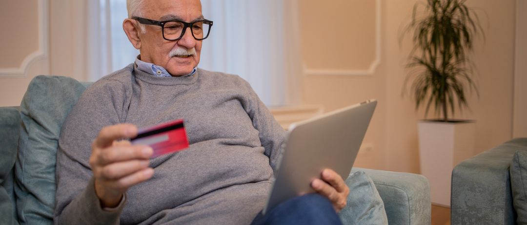 Ein älterer Mann mit Brille sitzt auf einem Sofa, hält eine Kreditkarte in der Hand und benutzt einen Laptop.