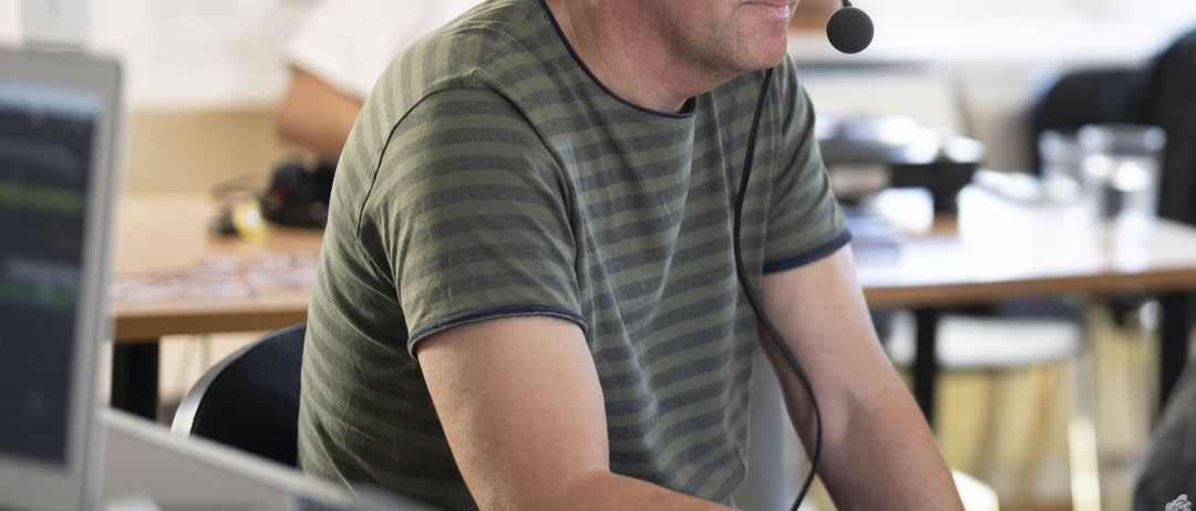 Ein Mann in einem grün gestreiften T-Shirt sitzt an einem Schreibtisch mit einem Mikrofon und Kopfhörern, arbeitet an einem Computer.