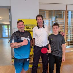 Drei Männer stehen lächelnd in einem Raum. Der Mann auf der linken Seite hat Tattoos am Arm. Der Mann in der Mitte hält eine Tischtennisschläger. Der Mann auf der rechten Seite trägt ein Nike-Shirt. Hinter ihnen ist ein gelbes Schild.