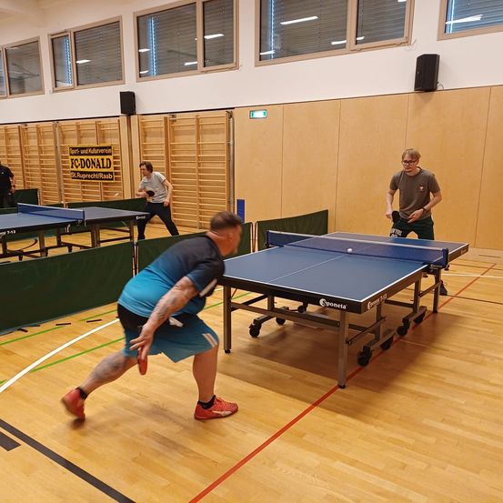 Drei Männer spielen Tischtennis in einer Halle mit einem blauen Tisch, Holzboden und Holzwänden. Ein Mann bereitet sich darauf vor, den Ball zu schlagen.
