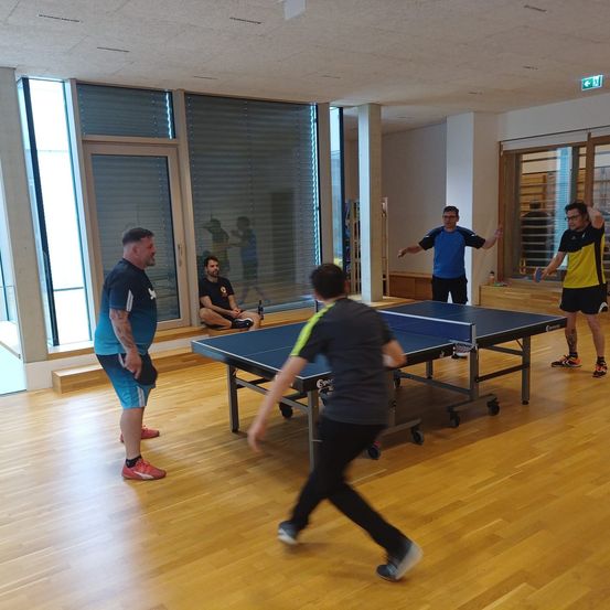 Fünf Männer spielen Tischtennis in einem Raum mit Holzboden und Glaswänden. Ein Mann in einem grauen Hemd läuft, um den Ball zu schlagen.