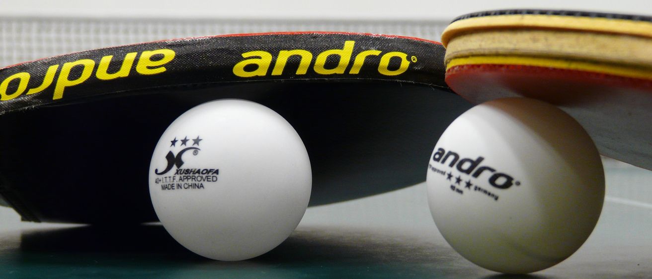 Zwei Tischtennisbälle, einer mit der Aufschrift 'andro' und der andere mit der Aufschrift 'KUSHAOFA ITTF APPROVED', liegen auf einem Tisch unter einem Tischtennisschläger mit der Aufschrift 'andro'.