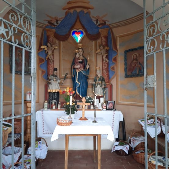 Eine kleine Kapelle mit einer Statue von Maria und Jesus, einem Kreuz und Blumen. Kerzen stehen auf dem Altar.