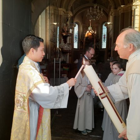 Zwei Priester halten brennende Kerzen in einer Kirche. Ein Mann und ein Mädchen helfen ihnen. Der Altar ist mit einem goldenen Kronleuchter geschmückt.