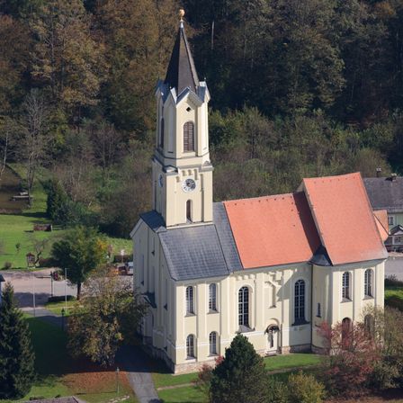 Eine Kirche mit Turm und rotem Dach steht auf einem grünen Feld, umgeben von Bäumen und Büschen. Die Kirche hat eine Uhr auf ihrem Turm.