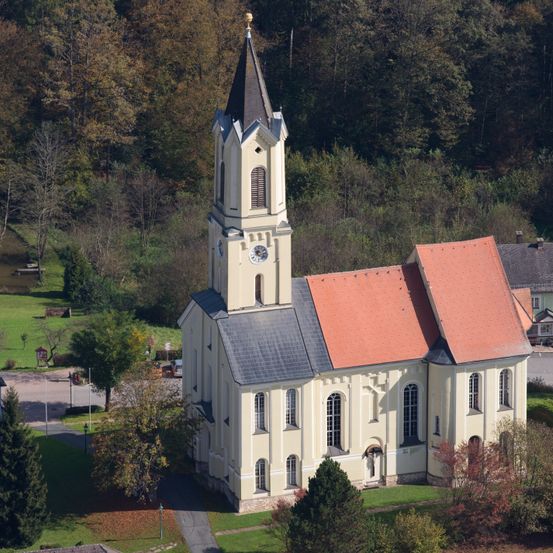 Eine Kirche mit Turm und rotem Dach steht auf einem grünen Feld, umgeben von Bäumen und Büschen. Die Kirche hat eine Uhr auf ihrem Turm.