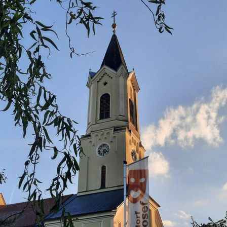 Ein Kirchturm mit einer Uhr und einem Banner für ein Theaterfestival. Der Turm ist mit einem Kreuz und einer goldenen Kugel geschmückt. Der Himmel ist teilweise bewölkt.