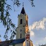 Ein Kirchturm mit einer Uhr und einem Banner für ein Theaterfestival. Der Turm ist mit einem Kreuz und einer goldenen Kugel geschmückt. Der Himmel ist teilweise bewölkt.