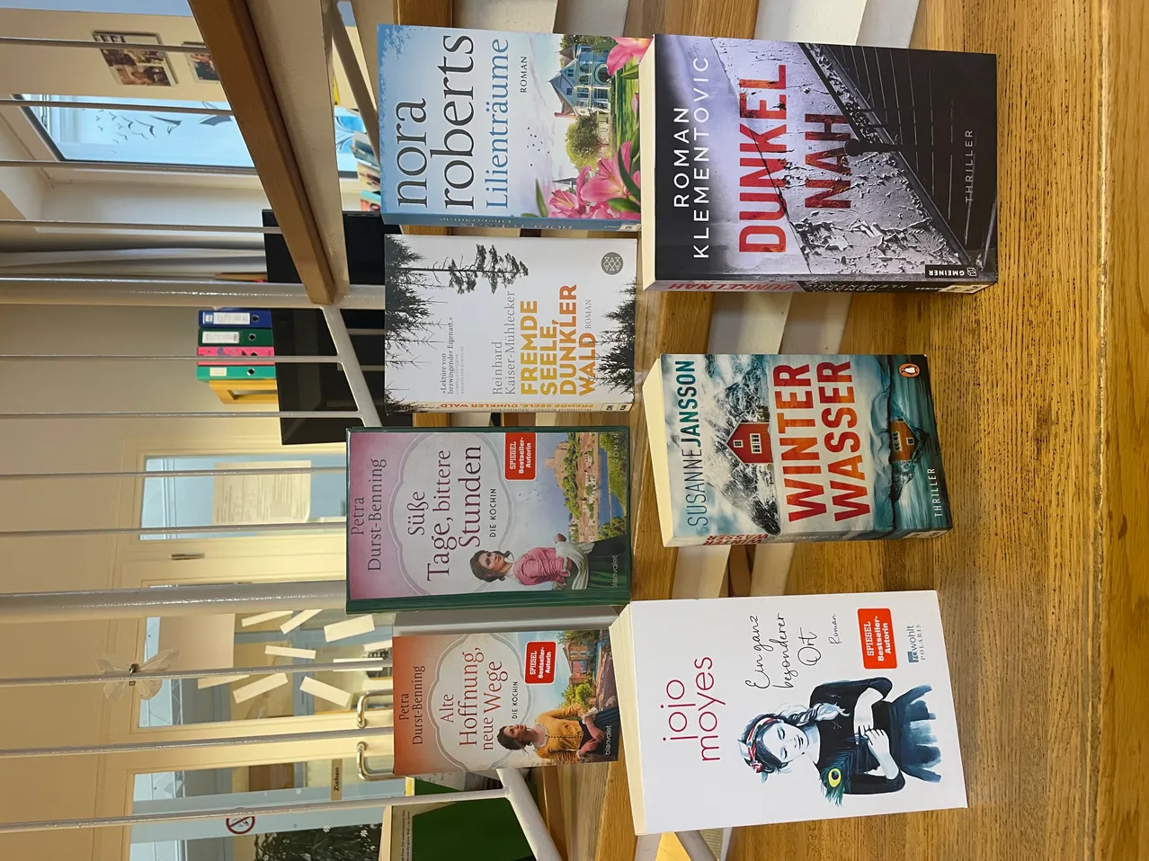 Mehrere Bücher sind auf einer Holzoberfläche gestapelt. Titel umfassen 'Lientrauma' von Nora Roberts, 'Fremde Senkeler' von Kerstin Gier, 'Dunkel' von Roman Vlcovic, 'Winter Wasser' von Susann Jansson, 'Tage, Stunden' von Petra Durst-Benning, 'Alte Wege neue Wege' von Petra Durst-Benning und 'ioyes' von Iio Moyes.