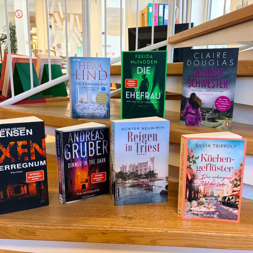 Sieben Bücher sind auf einem Holzregal ausgestellt. Ein Buch trägt den Titel 'Die Löwenmutter' von Hera Lind. Ein weiteres Buch heißt 'Die Ehefrau' von Freida McFadden. Das dritte Buch heißt 'Die geliebte Schwester' von Claire Douglas. Andere Bücher umfassen 'Reign in Trieste' von Gunter Neuwirth, 'Küchengeflüster' von Silvia Trippolt, 'Xen' von Jens Henrik Jensen und 'Dinner in the Dark' von Andreas Gruber.