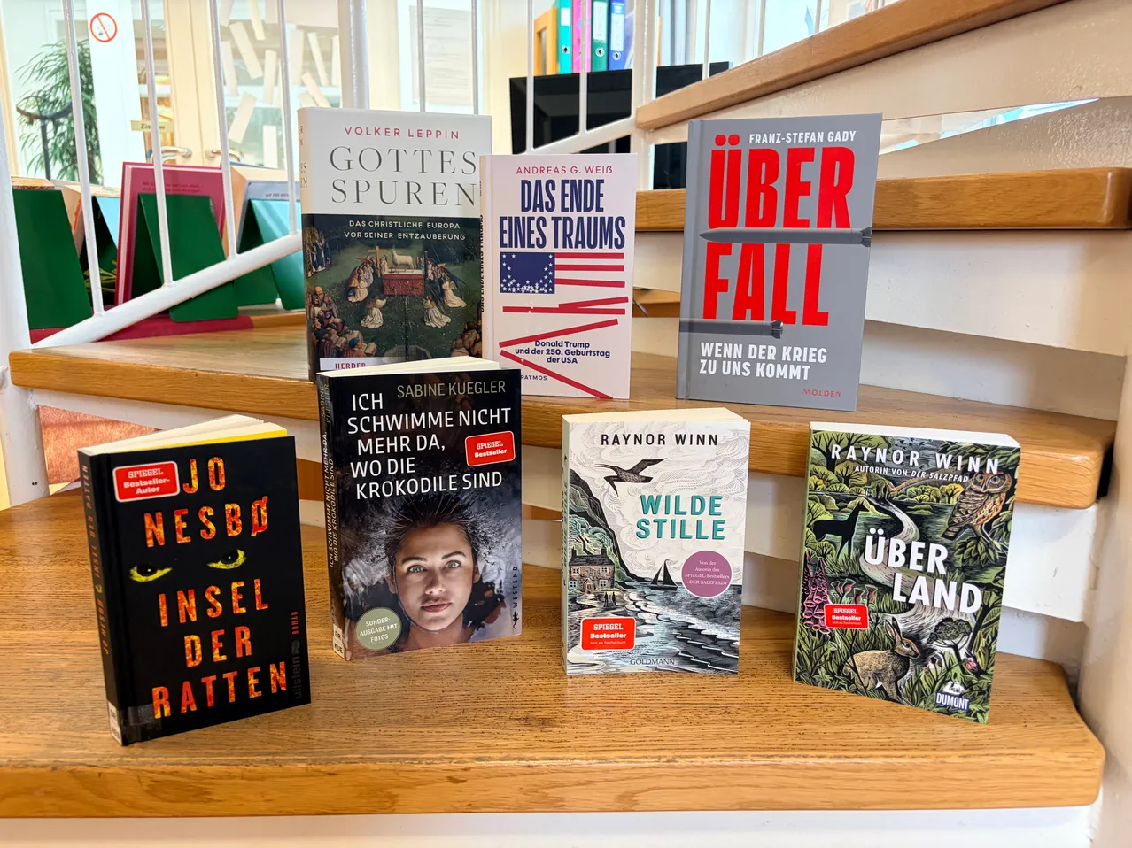 Ein Holzregal zeigt mehrere Bücher, darunter 'Gottes Spuren' von Volker Leppin, 'Das Ende eines Traums' von Andreas G. Weig, 'Über Fall' von Franz-Stefan Gady, 'Ich schwimme nicht mehr da, wo die Krokodile sind' von Sabine Kuegler, 'Wilde Stille' von Raynor Winn und 'Über Land' von Raynor Winn.