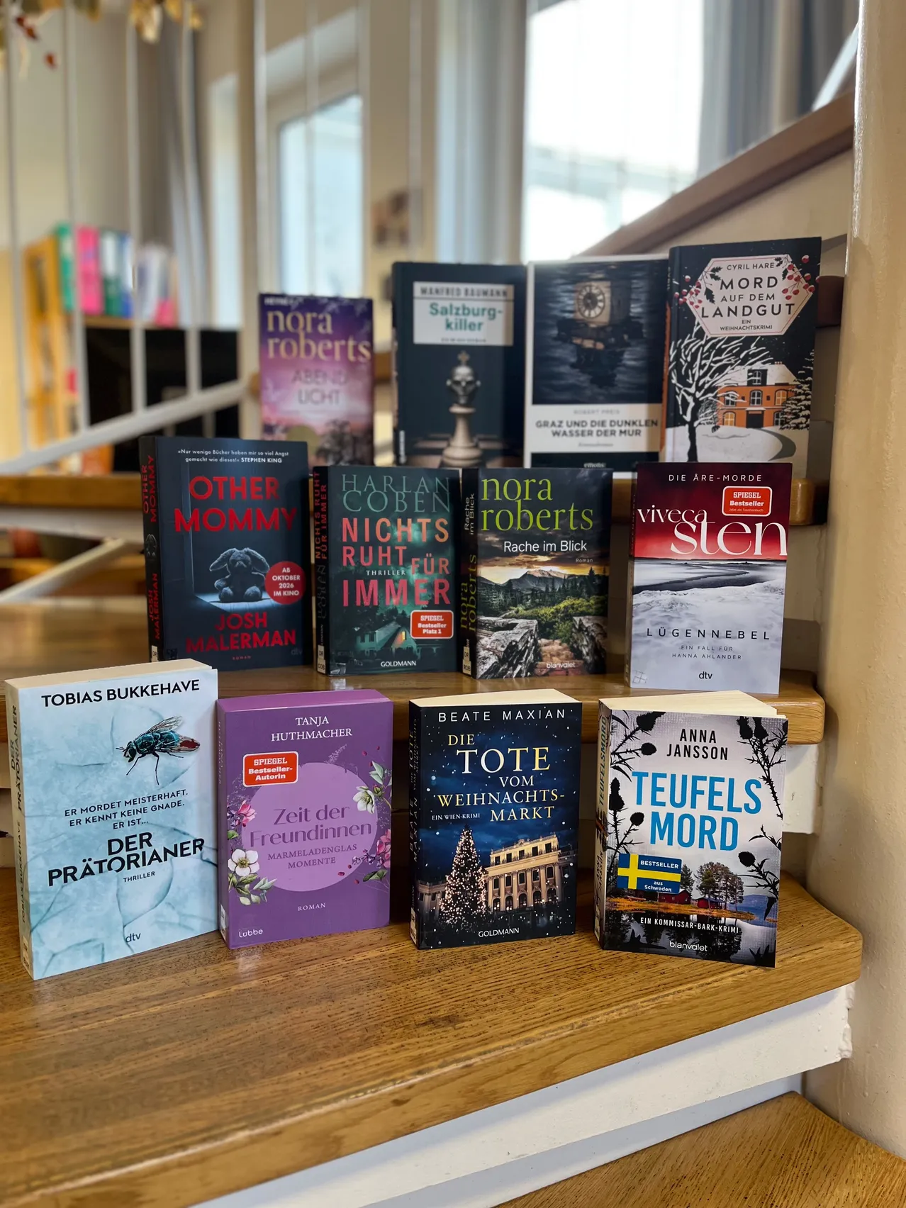 Ein Holzregal mit einer Sammlung von Büchern, darunter 'Other Mommy', 'Die Tote vom Weihnachtsmarkt', 'Teufel's Mord' und andere.