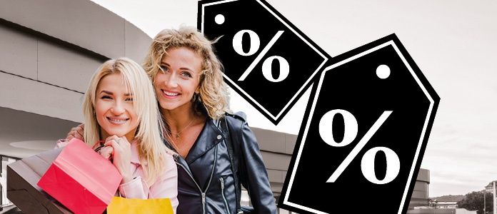 Zwei Frauen stehen mit Einkaufstüten vor einem Gebäude und lächeln. Das Gebäude hat ein modernes Design mit einem flachen Dach. Neben ihnen befinden sich Schilder mit Prozentsätzen.