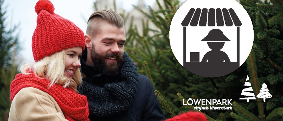 Ein Mann und eine Frau in Winterkleidung lächeln und schauen sich an der Tanne an. Im Hintergrund ist ein Logo für Löwenpark zu sehen.