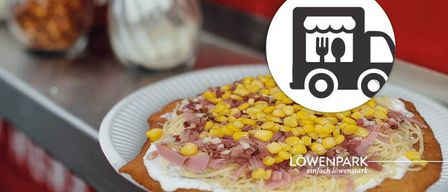 Ein weißer Teller hält ein Gericht mit Käse, Mais und Schinken. Ein Logo eines Lastwagens mit Gabel und Löffel ist auf dem Gericht. Eine rote Wand und Behälter sind im Hintergrund.