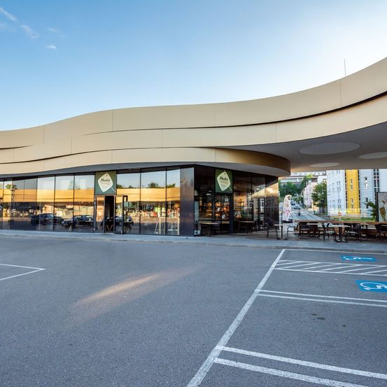 Außenansicht eines modernen Gebäudes mit Glasfenstern und einer offenen Vorderseite, mit Tischen und Stühlen draußen, unter einem klaren Himmel.