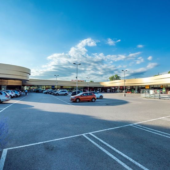 Ein leerer Parkplatz mit geparkten Autos und Straßenlaternen vor einem Einkaufszentrum an einem sonnigen Tag.