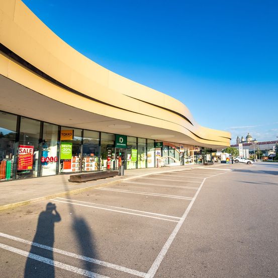 Ein großes Einkaufszentrum mit gebogenen gelben Wänden und Glasfenstern. Das Äußere ist sauber und gepflegt. Ein Auto ist vor dem Gebäude geparkt, und der Schatten einer Person fällt auf den Boden.