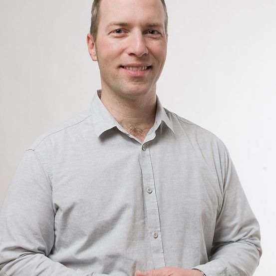 Ein Mann posiert für ein Foto mit einem hellgrauen Hemd und einer Uhr, er steht mit verschränkten Händen vor sich.