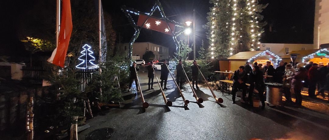 In der Nacht spielt eine Gruppe von Menschen lange Röhren vor einem geschmückten Baum. Eine beleuchtete Flagge ist links. Ein Feuer brennt rechts.