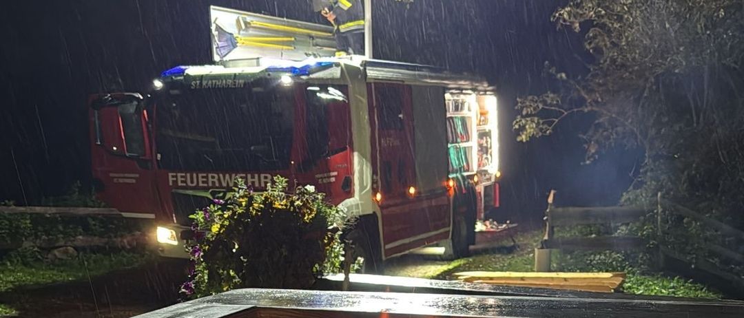 Ein Feuerwehrfahrzeug mit leuchtenden Lichtern steht im Regen, mit einem brennenden Feuerkorb in der Nähe.
