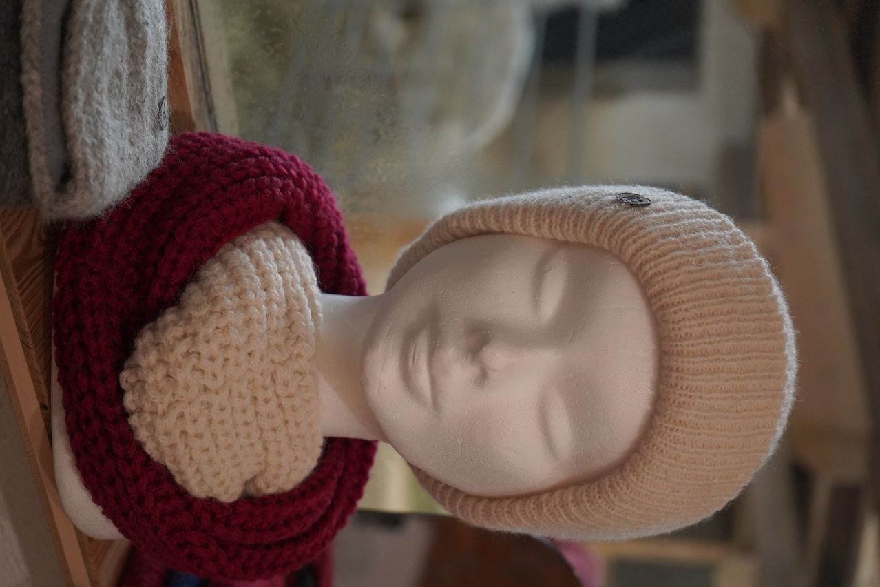A close-up of a mannequin's head with a knitted beanie and scarf, in front of a blurry glass window.