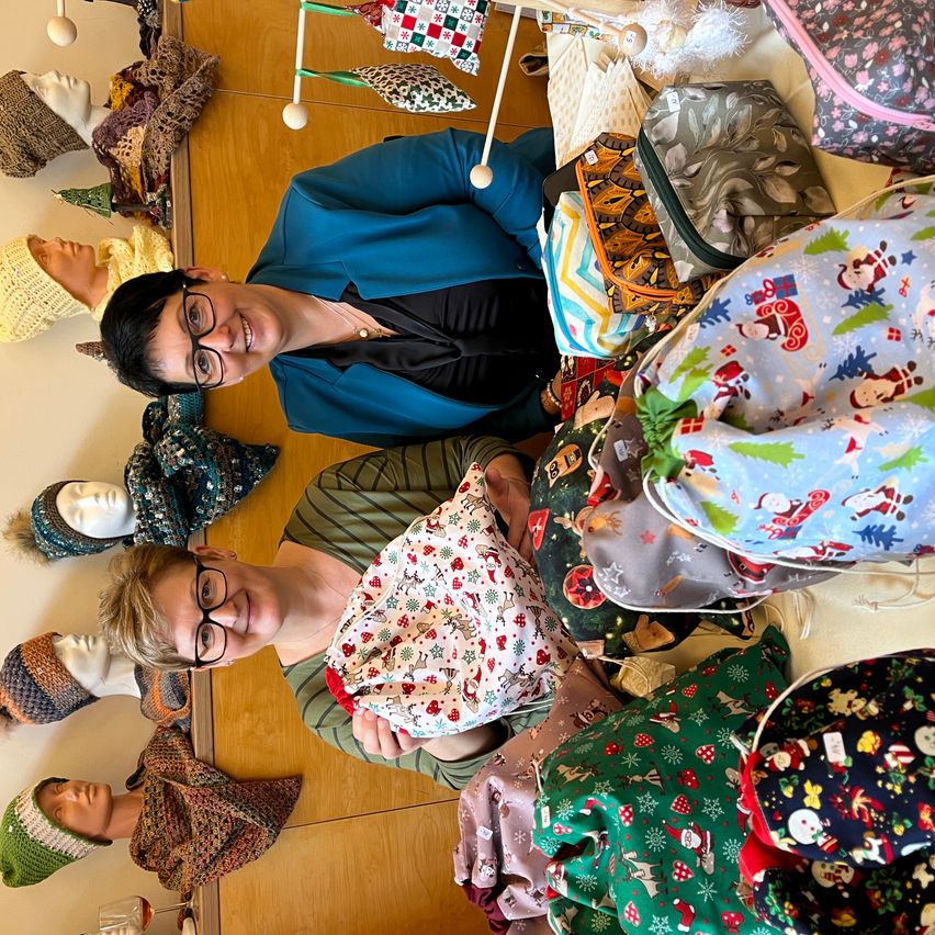 A group of people are gathered around a table, surrounded by various handmade Christmas items. Two women are smiling and holding Christmas-themed bags. Behind them, mannequin heads with knitted hats and scarves are on display.