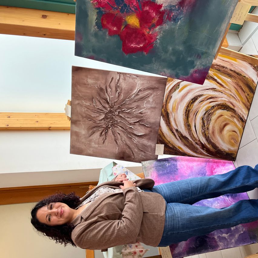 A woman is lying on the floor, smiling, in front of several paintings. Above her, three paintings hang on the wall.