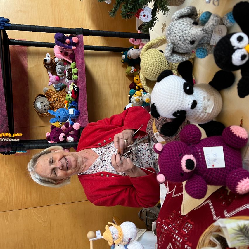 An older woman with gray hair crochets a purple bear with pink details. She is surrounded by a variety of crocheted animals, including a panda, elephant, and penguin. The background shows a wooden wall with a shelf holding various stuffed toys.
