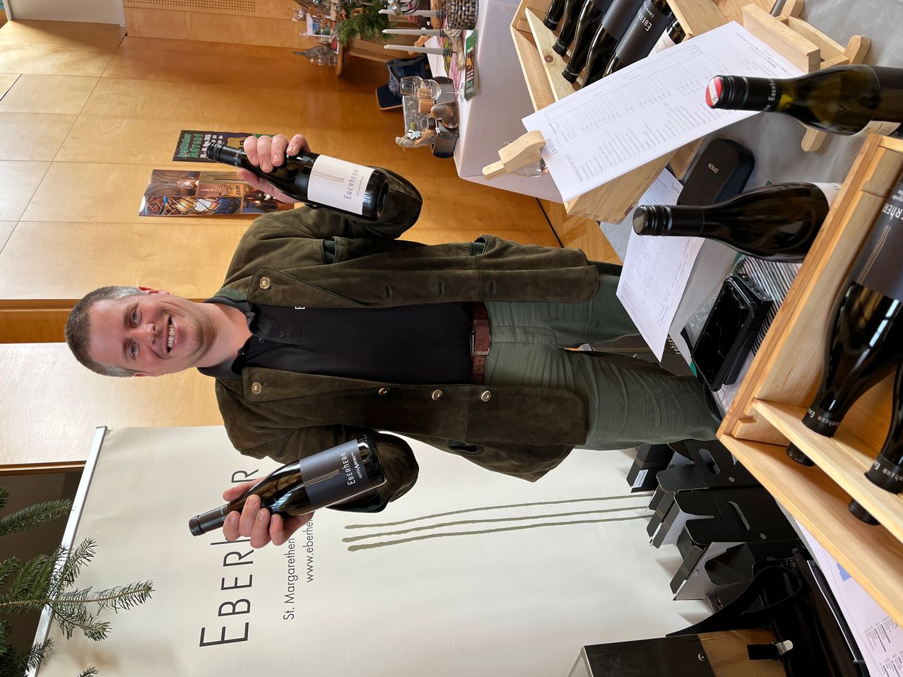 A man stands at a wine display, holding a bottle in each hand, smiling, with several other bottles on the table and a banner reading 'EBERLE' behind him.