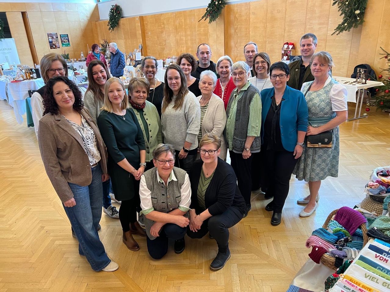 A group of women and men are smiling and posing for a photo inside a room with wooden floors.