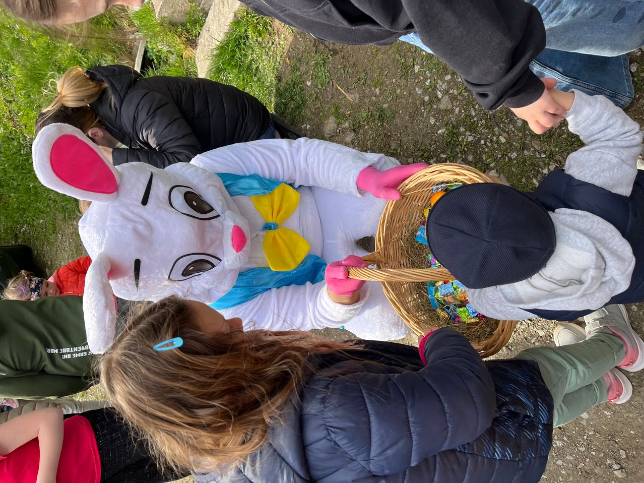 A group of children gather around an Easter Bunny mascot who is holding a basket filled with candy. A girl in a blue jacket leans in to look.