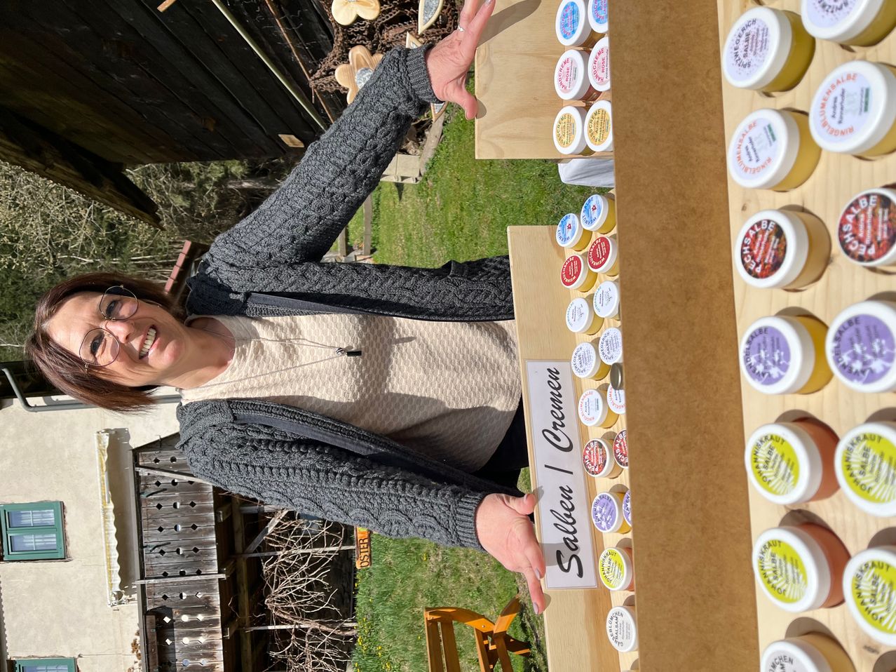A woman stands behind a display of jars with cream, smiling and pointing to a sign that reads 'Sallei & Cremen'. She is dressed in a gray sweater and holds a wooden display with a label.