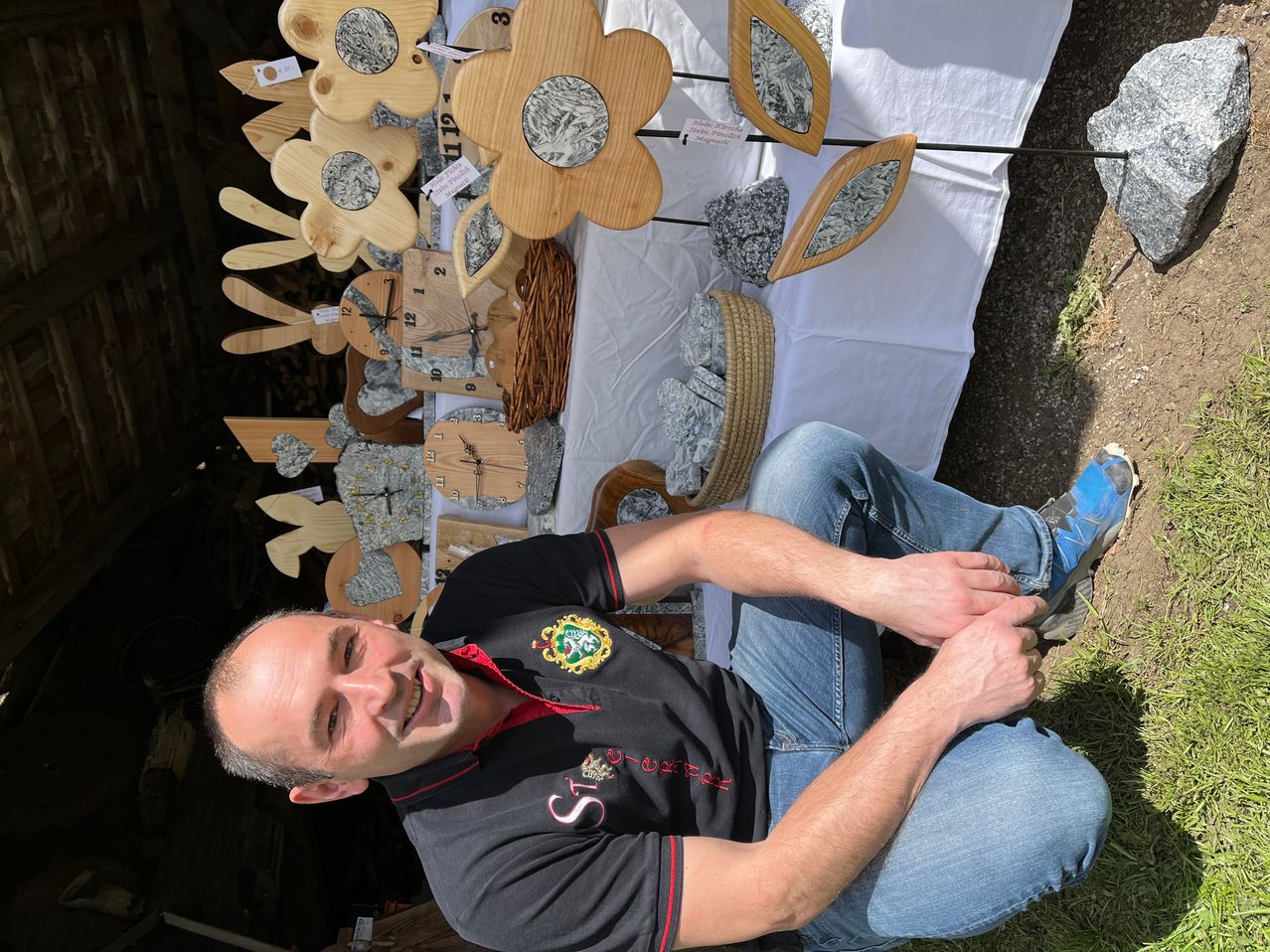 A man is sitting on the ground, smiling, with wooden crafts and stone pieces behind him. The wooden crafts include flowers and clocks.