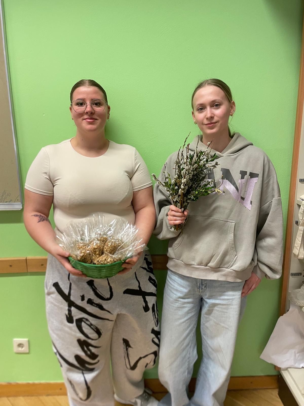 Zwei Frauen stehen gegen eine grüne Wand, eine hält einen grünen Korb mit einer Blumenarrangement und die andere hält einen Strauß.