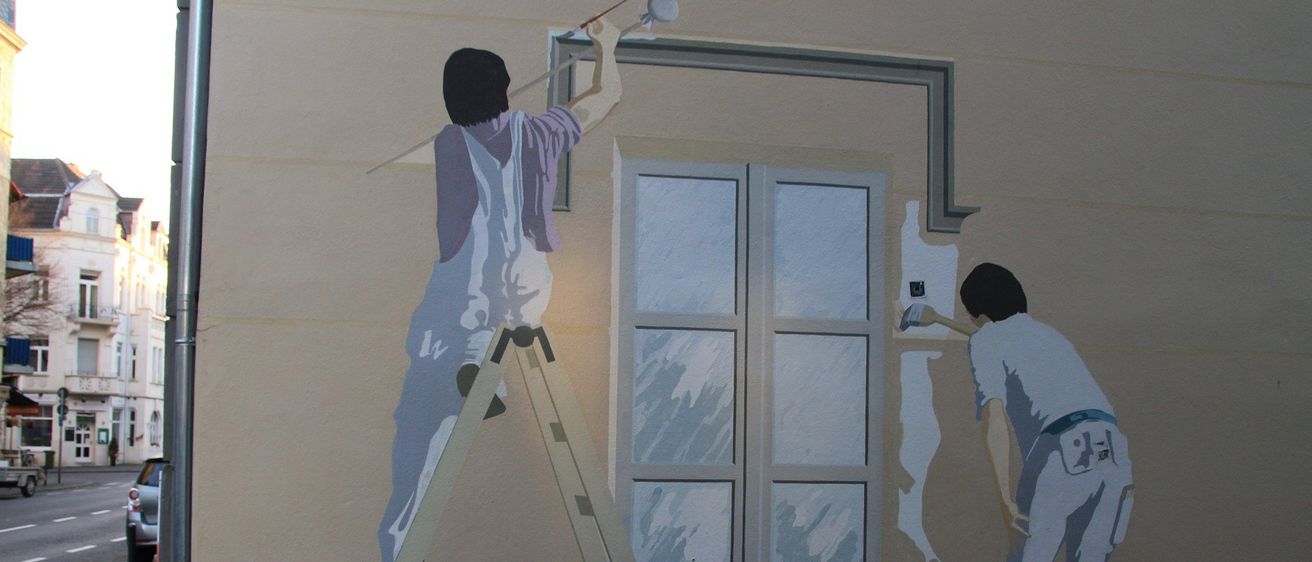 Ein Wandbild zeigt zwei Männer, die ein Gebäude malen. Einer steht auf einer Leiter und malt die Wand, während der andere auf der Fensterbank steht und den Fensterrahmen malt.