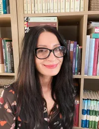 Eine Frau mit langen schwarzen Haaren und einer Brille steht vor einem Bücherregal, das mit Büchern gefüllt ist.