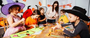Mehrere Kinder in Halloween-Kostümen basteln an einem Tisch, angeleitet von einer Frau mit Brille. Der Raum ist mit Halloween-Dekorationen geschmückt.