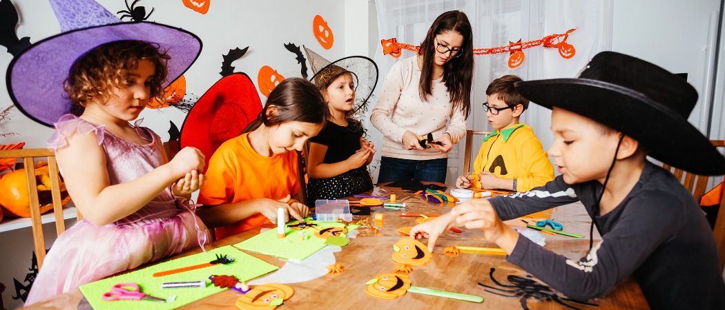 Mehrere Kinder in Halloween-Kostümen basteln an einem Tisch, angeleitet von einer Frau mit Brille. Der Raum ist mit Halloween-Dekorationen geschmückt.