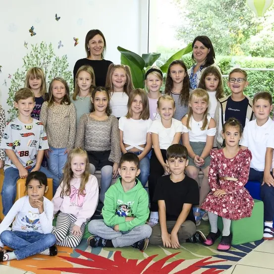 Ein Klassenfoto von Kindern, Lehrern und Pflanzen. Die Kinder und Lehrer lächeln und posieren für die Kamera. Die Kinder sitzen auf dem Boden, während die Lehrer hinter ihnen stehen.