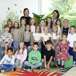 Ein Klassenfoto von Kindern, Lehrern und Pflanzen. Die Kinder und Lehrer lächeln und posieren für die Kamera. Die Kinder sitzen auf dem Boden, während die Lehrer hinter ihnen stehen.