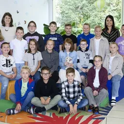 Eine Gruppe von Kindern und zwei Frauen posiert für ein Foto, lächelt und sitzt auf bunten Kissen. Die Kinder tragen verschiedene Kleidungsstücke, und der Raum hat ein Glasfenster, das Bäume draußen zeigt.
