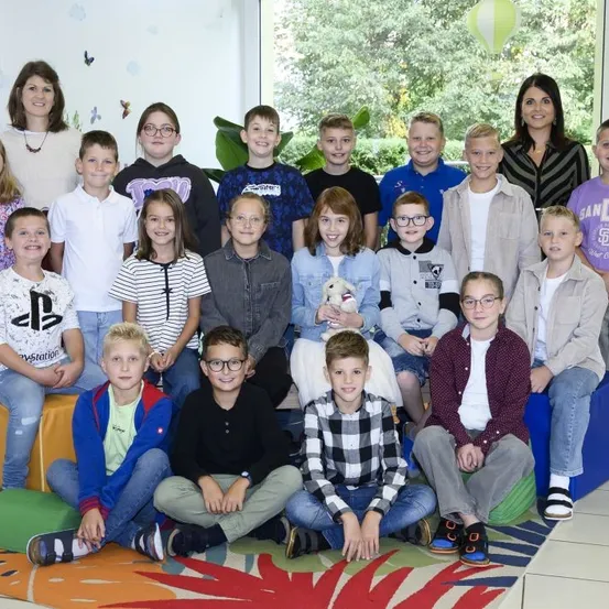 Eine Gruppe von Kindern und zwei Frauen posiert für ein Foto, lächelt und sitzt auf bunten Kissen. Die Kinder tragen verschiedene Kleidungsstücke, und der Raum hat ein Glasfenster, das Bäume draußen zeigt.