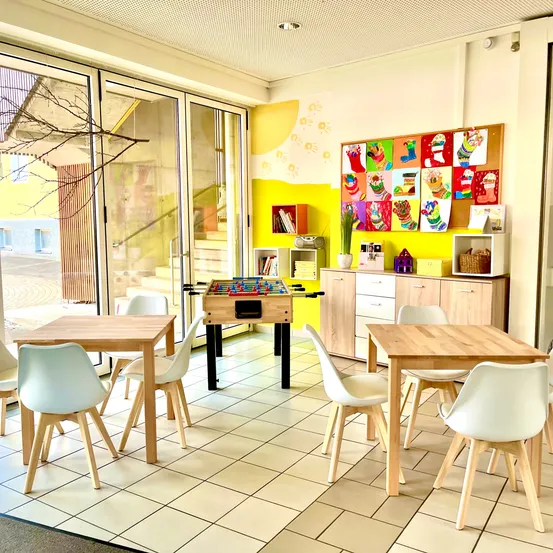 Ein heller Raum mit gelben Wänden, der zwei Holztische, weiße Stühle und einen Tischkicker bietet. Es gibt eine Pinnwand mit bunten Kunstwerken und Schränke mit Mikrowelle und Korb. Glastüren führen in einen Flur.